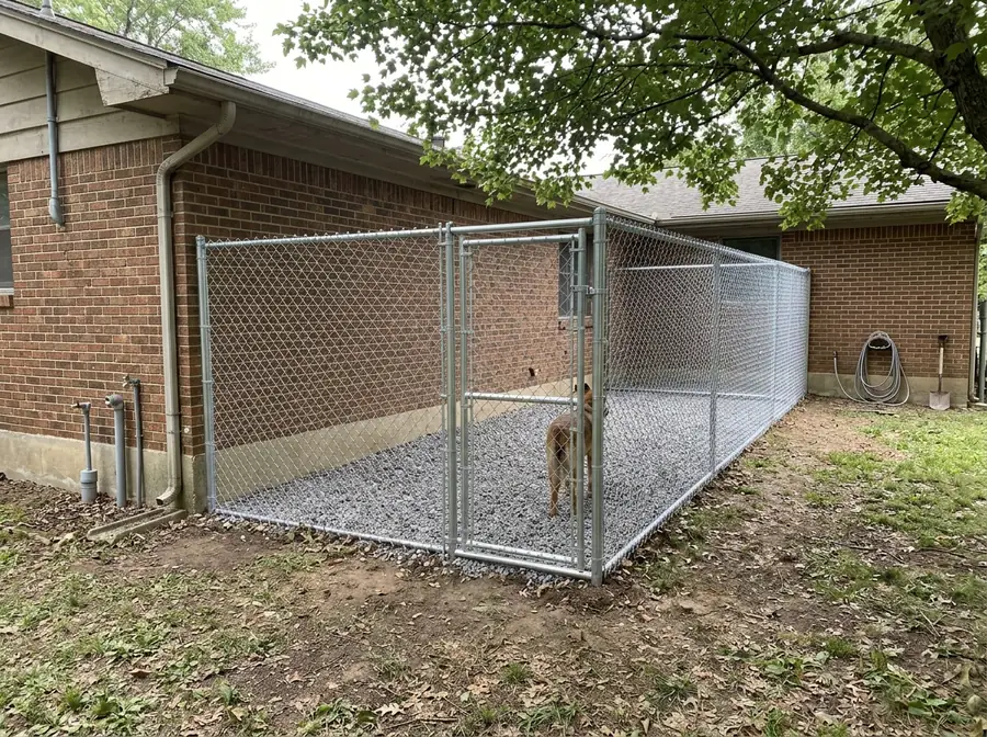 A galvanized chain link dog run with a gate along the side of a house on a gravel base