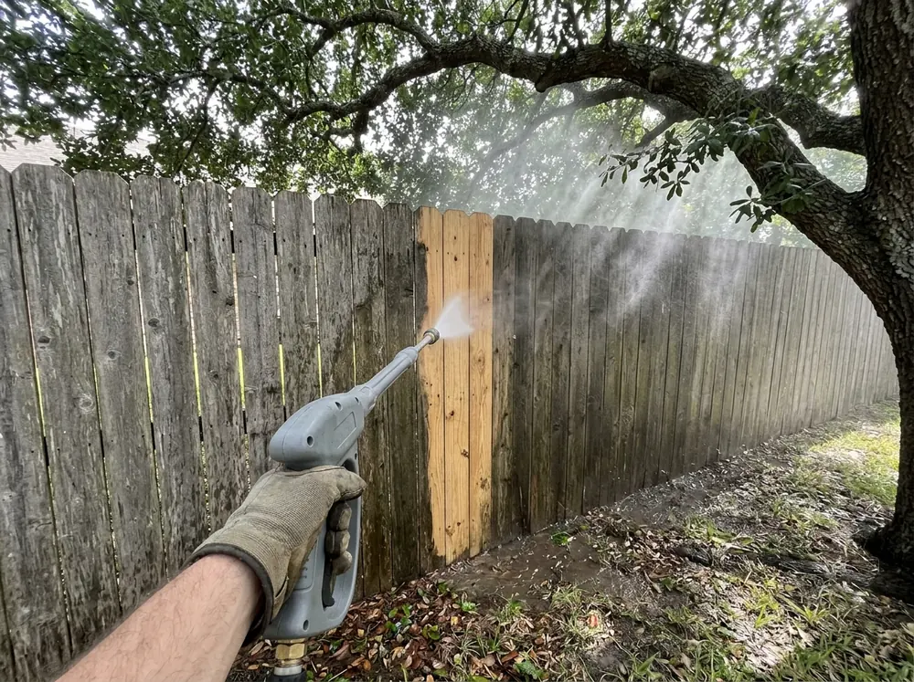 A pressure washer wand stripping green mildew from a gray wood fence, leaving a clean stripe